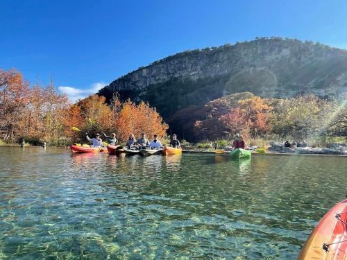 A group of people kayaking on a clear lake surrounded by autumn foliage and a mountainous backdrop.