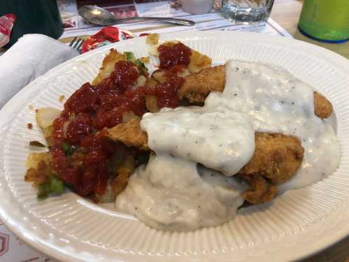 A plate of fried chicken topped with creamy gravy, served with hash browns and ketchup on the side.