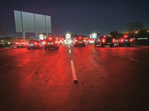 Cars lined up at night, illuminated by red brake lights, with a large blank billboard in the background.