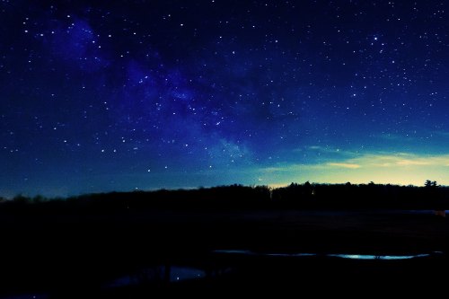 A starry night sky with a visible Milky Way, silhouetted trees, and a calm water reflection in the foreground.