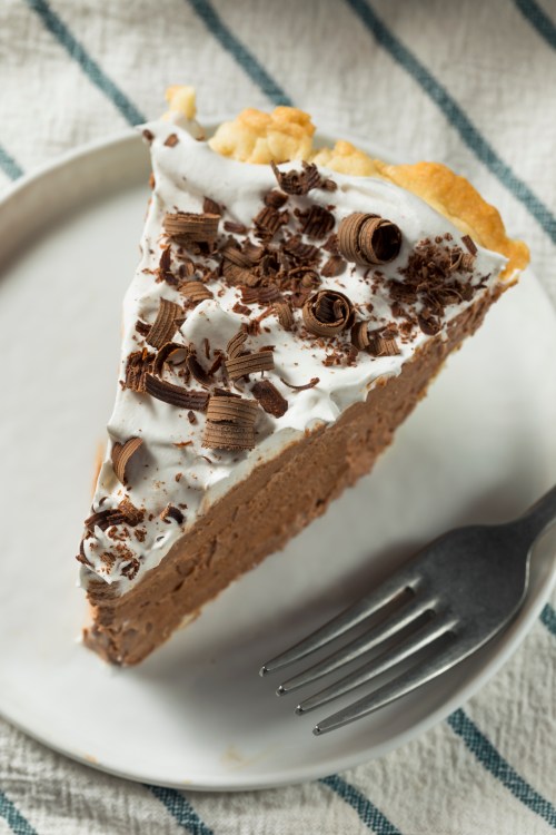 A slice of chocolate cream pie topped with whipped cream and chocolate shavings on a white plate with a fork.