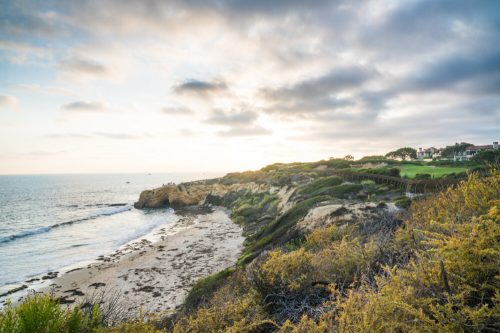 Coastal landscape at sunset, featuring rocky cliffs, sandy beach, and gentle waves under a cloudy sky.