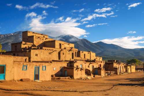 Historic adobe buildings under a blue sky with mountains in the background, showcasing a desert landscape.