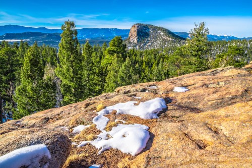 A rocky landscape with patches of snow, surrounded by evergreen trees and distant mountains under a clear blue sky.