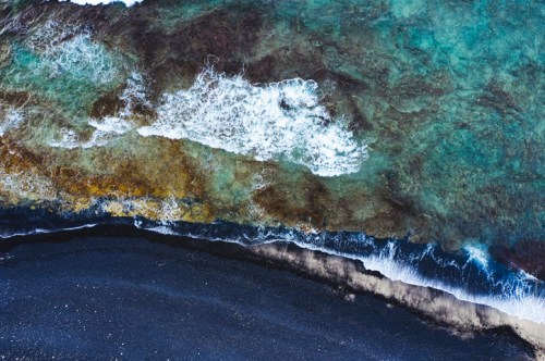 Aerial view of a coastline with turquoise water, rocky shore, and black sand beach. Waves gently crash along the edge.