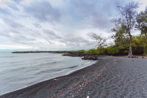 A serene black sand beach with gentle waves, surrounded by lush greenery and a cloudy sky.
