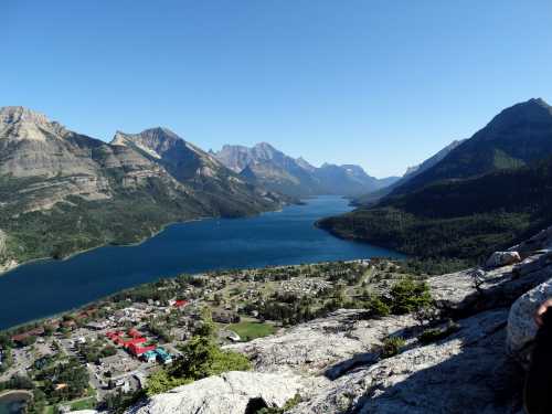 A scenic view of a lake surrounded by mountains, with a small town visible along the shoreline under a clear blue sky.