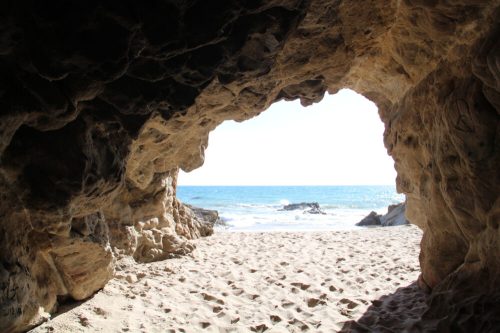 View from a cave opening, revealing a sandy beach and ocean waves under a bright sky.
