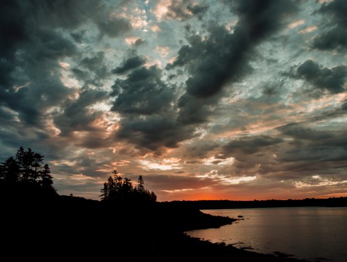 A serene sunset over a calm lake, with dramatic clouds and silhouettes of trees along the shoreline.