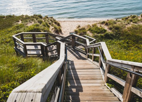 Wooden stairs lead down to a sandy beach, surrounded by green grass and calm blue water.