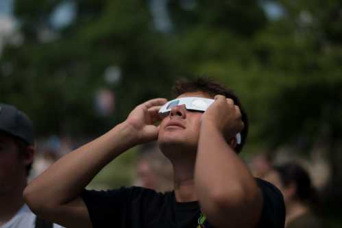 A young man wearing eclipse glasses looks up, shielding his eyes, with trees in the background.