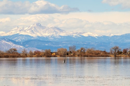 A serene lake reflects snowy mountains under a blue sky, with trees and a distant house along the shore.