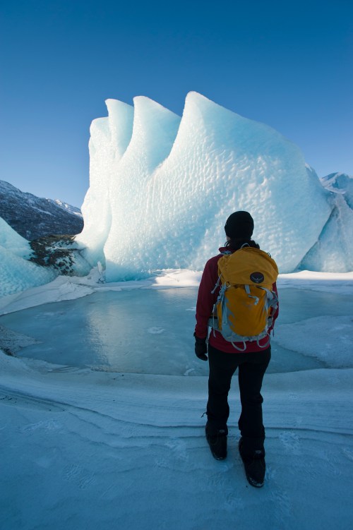 A person stands on icy terrain, gazing at a large, jagged ice formation under a clear blue sky.