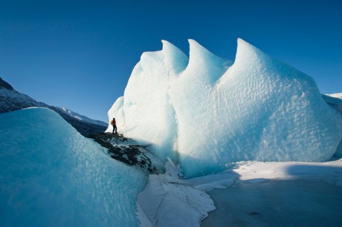 A person stands on a rocky surface near towering blue ice formations under a clear blue sky.