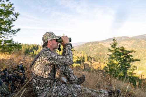 A hunter in camouflage sits on a hillside, using binoculars to scan the landscape for wildlife.