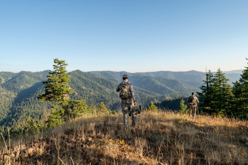 Two hunters in camouflage gear walking on a hillside, overlooking a vast mountainous landscape under a clear sky.