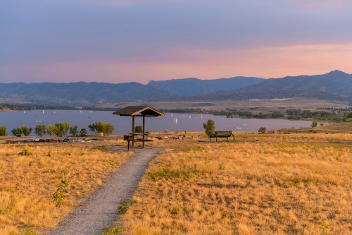 A scenic view of a lake surrounded by mountains, with a path leading to a gazebo and benches under a colorful sky.