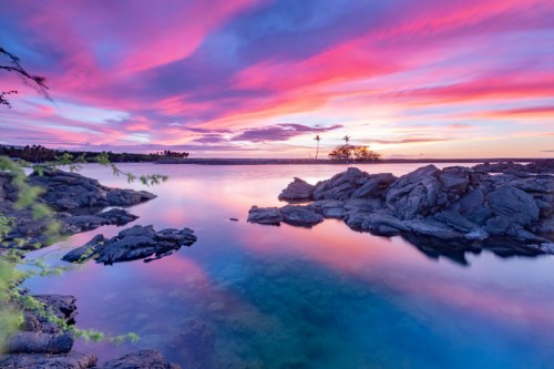 A serene landscape featuring a colorful sunset over calm waters, with rocky shores and silhouetted palm trees.