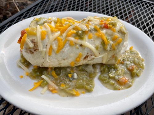 A large burrito topped with melted cheese and green sauce, served on a plate with a side of sauce and toppings.