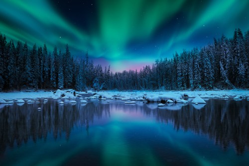 A serene winter landscape with a river reflecting vibrant green and blue northern lights above a snowy forest.