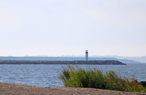 A lighthouse stands on a rocky shore, overlooking a calm sea with distant hills in the background.