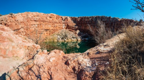 A rocky canyon with a small, green pond surrounded by dry vegetation under a clear blue sky.