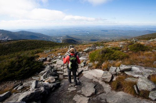 A hiker in a red jacket walks down a rocky trail, surrounded by mountains and a vast landscape under a blue sky.