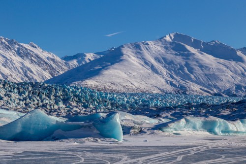 A snowy landscape featuring blue ice formations and mountains under a clear blue sky.