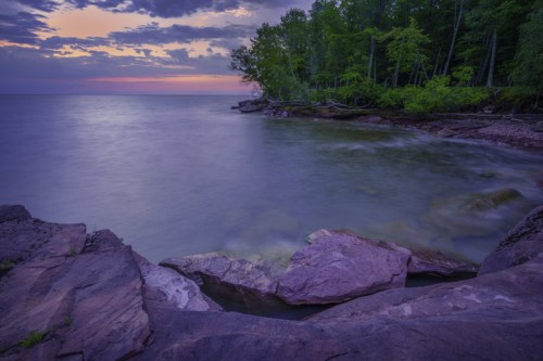 Serene lakeside view at dusk, with smooth water, rocky shore, and lush trees under a colorful sky.