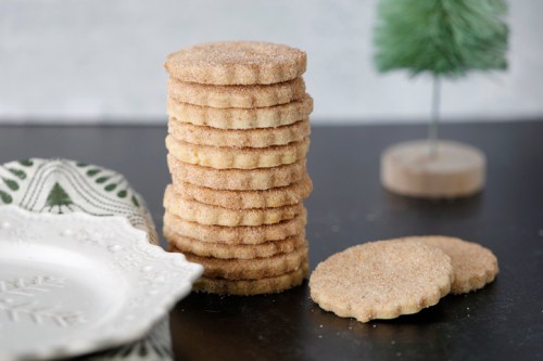 A stack of sugar cookies sits on a dark table, with a decorative plate and a small green tree in the background.