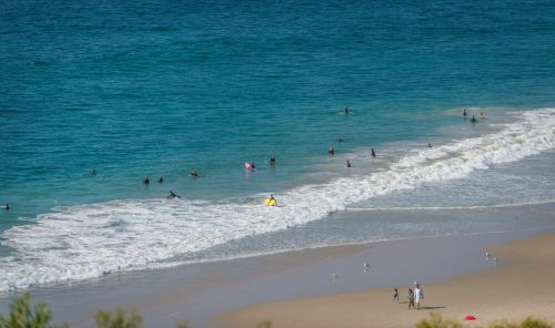 A sunny beach scene with people swimming, playing, and relaxing by the waves under a clear blue sky.