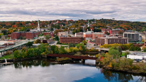 Aerial view of a city with colorful autumn trees, buildings, and a river reflecting the cloudy sky.