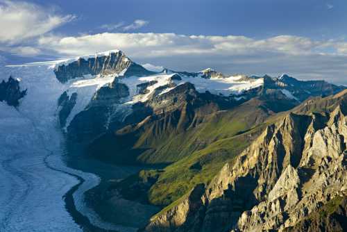 A stunning mountain landscape featuring glaciers, rugged peaks, and lush green valleys under a blue sky with clouds.