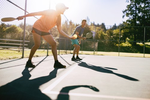 Two people playing pickleball on a sunny court, casting long shadows on the ground.