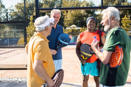 Four older men chatting and laughing on a padel court, holding paddles and a water bottle.