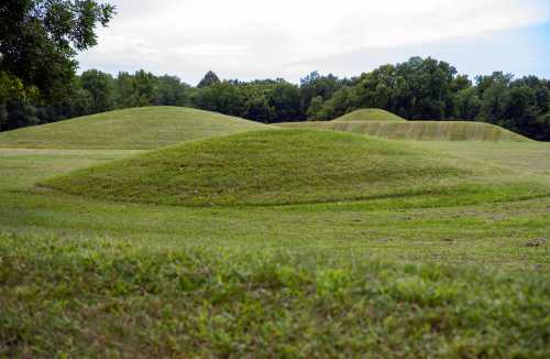 Three grassy mounds rise gently in a green landscape, surrounded by trees under a cloudy sky.