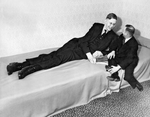 A tall man reclines on a bed while a shorter man sits beside him, both dressed in formal attire.