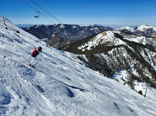 A skier descends a snowy mountain slope with a clear blue sky and distant peaks in the background.