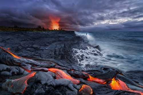 Lava flows into the ocean at dusk, with dramatic clouds and a distant eruption illuminating the sky.