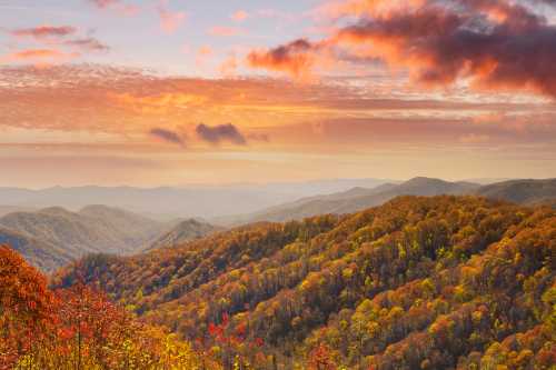 A vibrant sunset over rolling mountains, showcasing autumn foliage in shades of orange, red, and yellow.