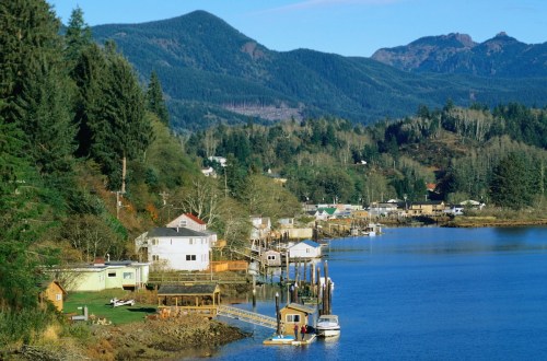 A scenic view of a riverside town with houses, docks, and mountains in the background under a clear blue sky.