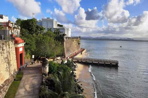 Scenic view of a coastal area with a boardwalk, lush greenery, and historic walls under a partly cloudy sky.