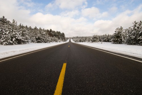 A snow-covered road stretches into the distance, flanked by trees under a cloudy sky.