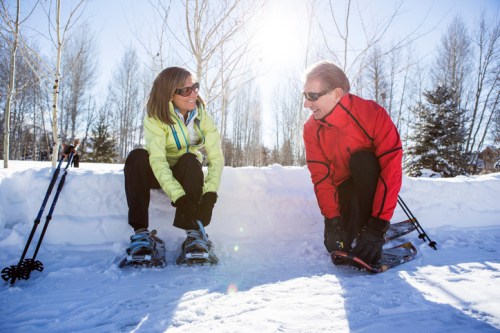 Two people in winter gear sit on a snowbank, putting on snowshoes, with trees and sunlight in the background.
