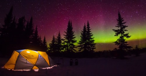 A glowing tent in a snowy landscape under a vibrant aurora and starry night sky, surrounded by tall trees.
