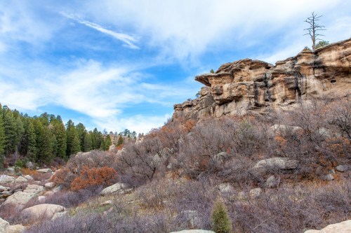 Rocky cliffs rise above a landscape of sparse trees and shrubs under a blue sky with scattered clouds.