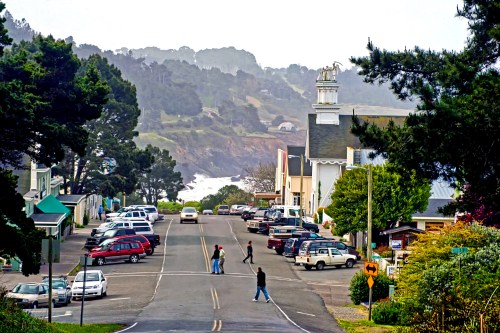 A coastal town street with parked cars, pedestrians crossing, and a church in the background amidst lush greenery.