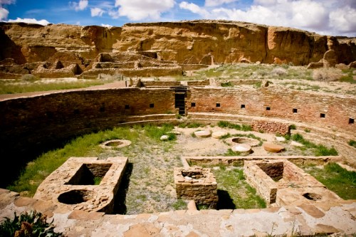 Ancient stone ruins surrounded by grass and cliffs under a blue sky with clouds.