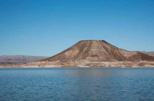 A serene landscape featuring a conical mountain rising above a calm blue lake under a clear blue sky.