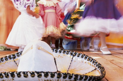 A blurred scene of dancers in colorful dresses, with a sombrero in focus on a wooden floor.